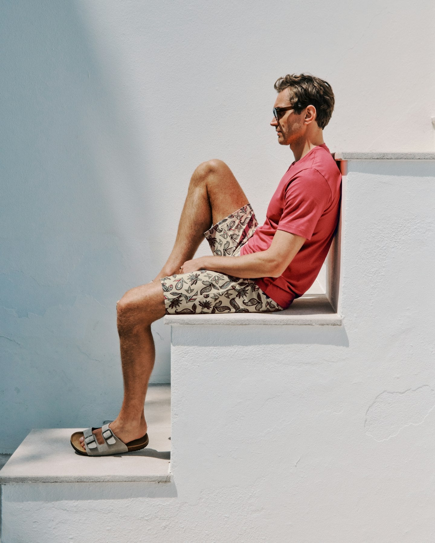 Man sitting on a white ledge wearing a red shirt and patterned shorts.