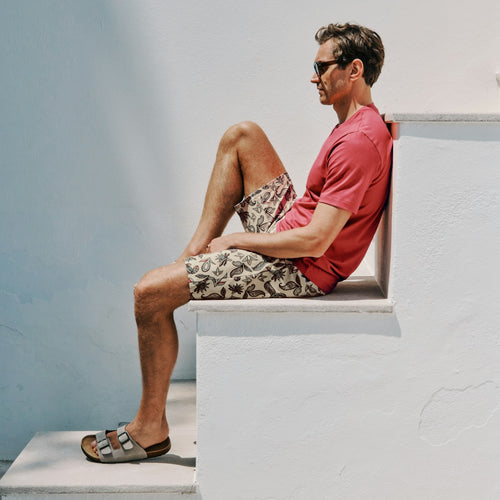 Man sitting on a white ledge wearing a red shirt and patterned shorts.