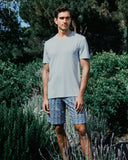 Man wearing a light gray t-shirt and patterned shorts standing in a garden with greenery and lavender.