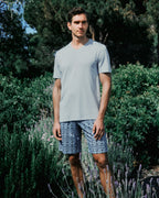 Man wearing a light gray t-shirt and patterned shorts standing in a garden with greenery and lavender.