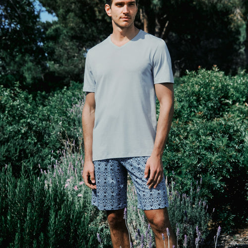 Man wearing a light gray t-shirt and patterned shorts standing in a garden with greenery and lavender.