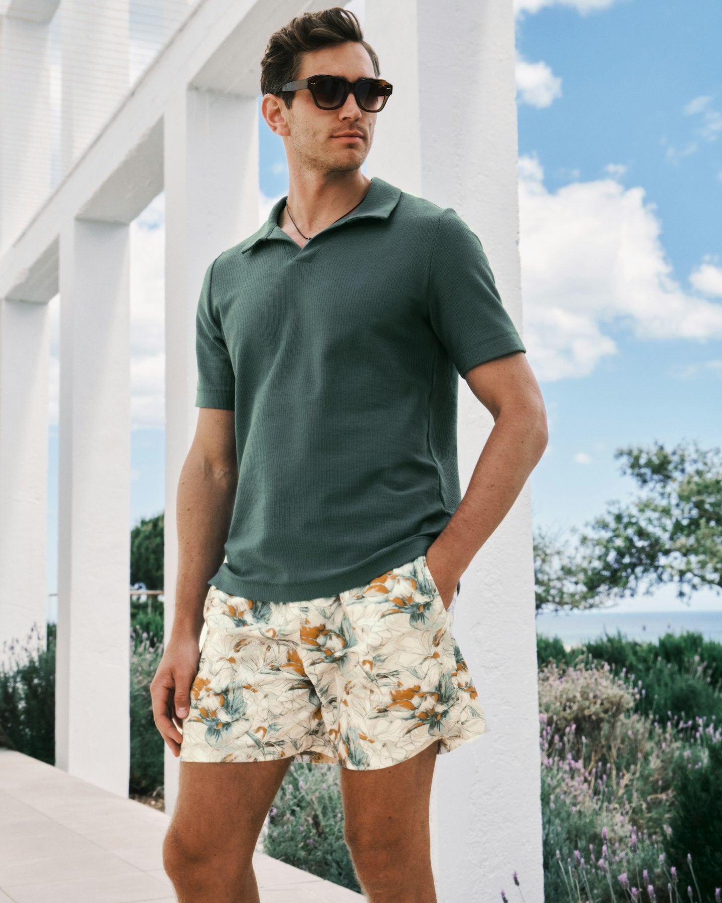 Man wearing a green polo shirt and floral shorts standing outdoors with white columns and blue sky in the background.
