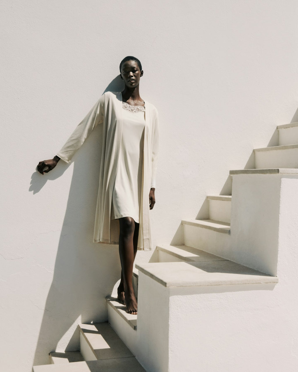 Woman in a long beige coat standing on a white staircase against a white wall