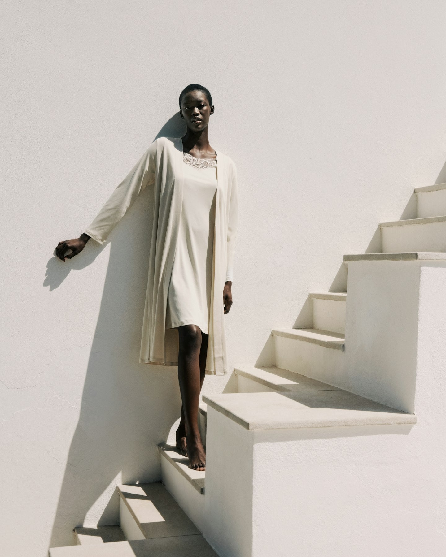 Woman in a long beige coat standing on a white staircase against a white wall