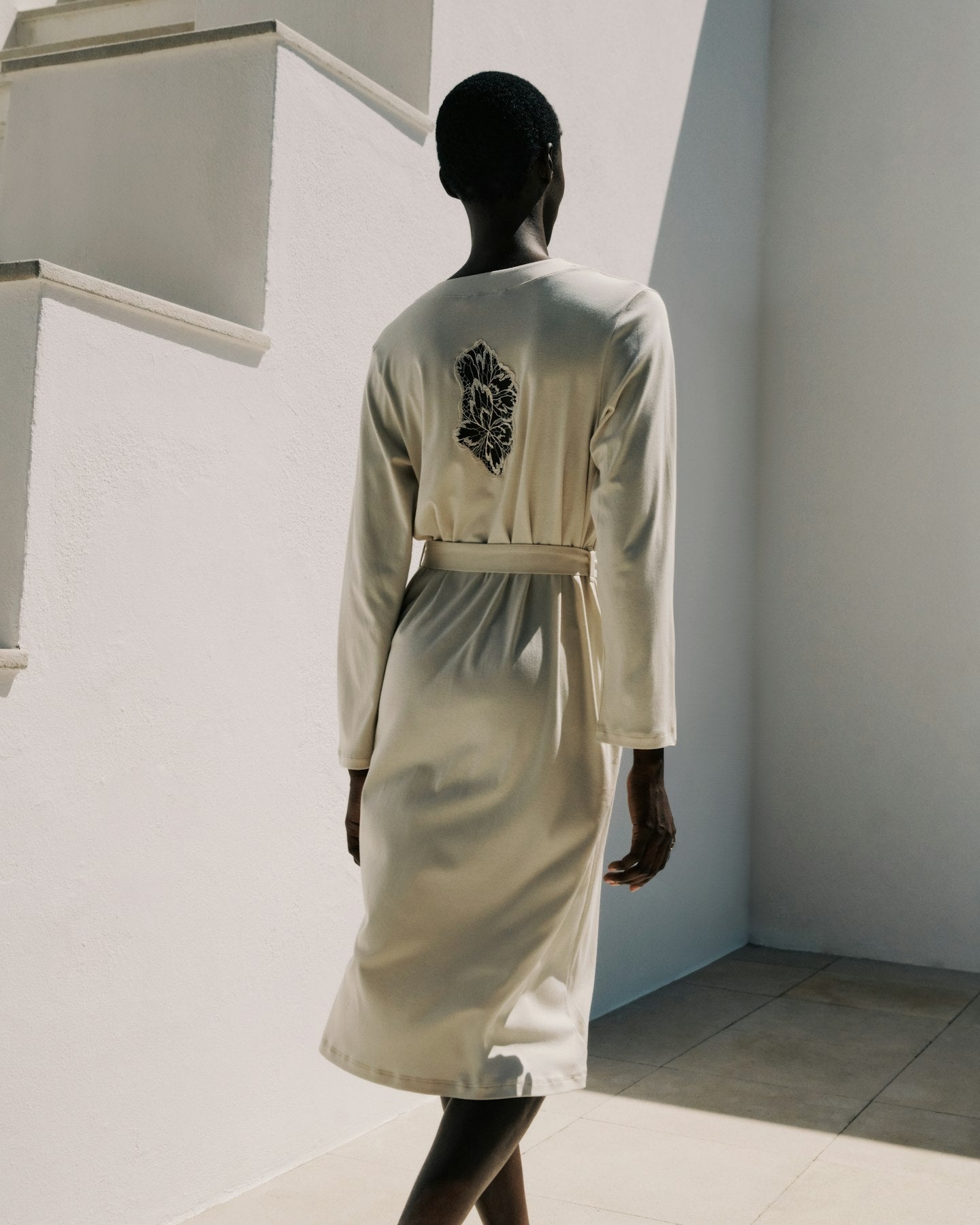Mannequin wearing a light-colored robe with a decorative back design against a white wall.