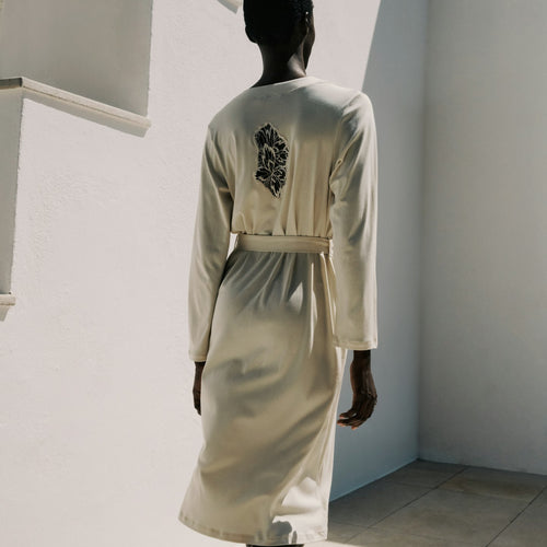 Mannequin wearing a light-colored robe with a decorative back design against a white wall.