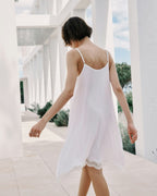 Woman in a white slip dress walking in a sunlit outdoor setting with columns and greenery.