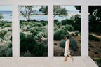 Woman walking on a path with a scenic view of trees and water through large windows.