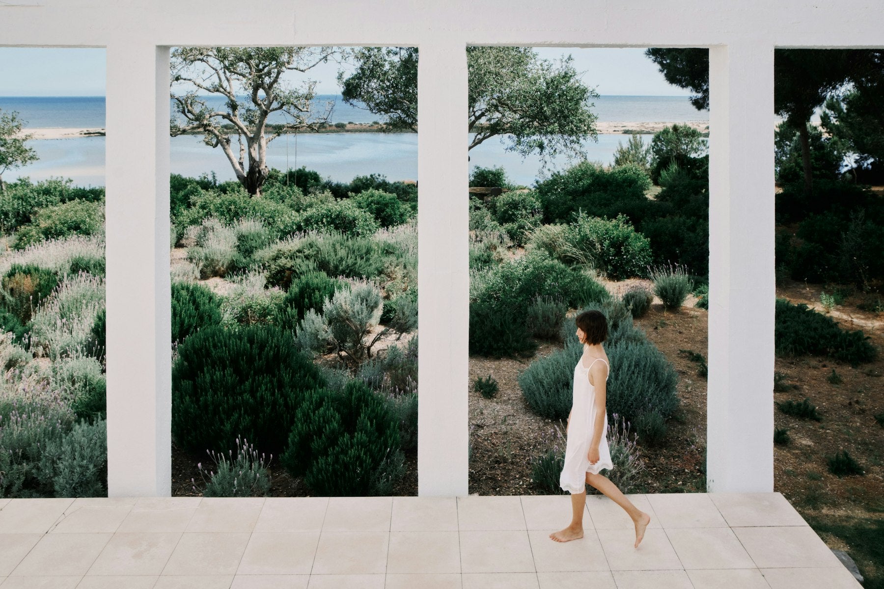Woman walking on a path with a scenic view of trees and water through large windows.