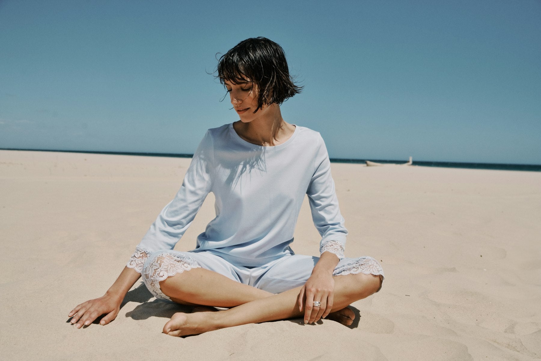 Woman sitting on a sandy beach wearing a light blue long-sleeve shirt and shorts.