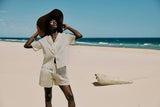 Person wearing a wide-brimmed hat matching cotton shirt and shorts on a sandy beach with clear blue sky and ocean.