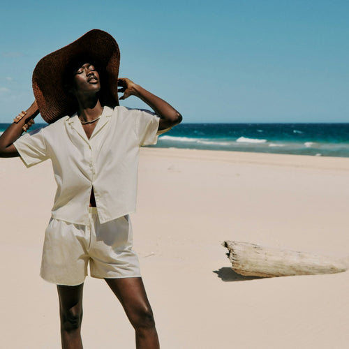 Person wearing a wide-brimmed hat matching cotton shirt and shorts on a sandy beach with clear blue sky and ocean.