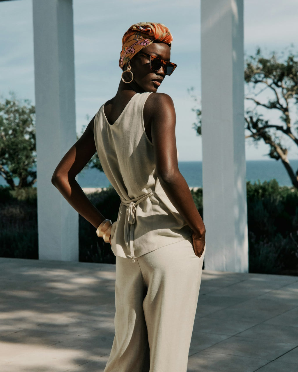 Woman in a beige outfit with a headscarf and sunglasses standing outdoors with ocean view.