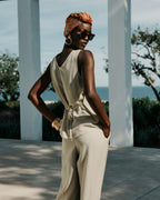 Woman in a beige outfit with a headscarf and sunglasses standing outdoors with ocean view.