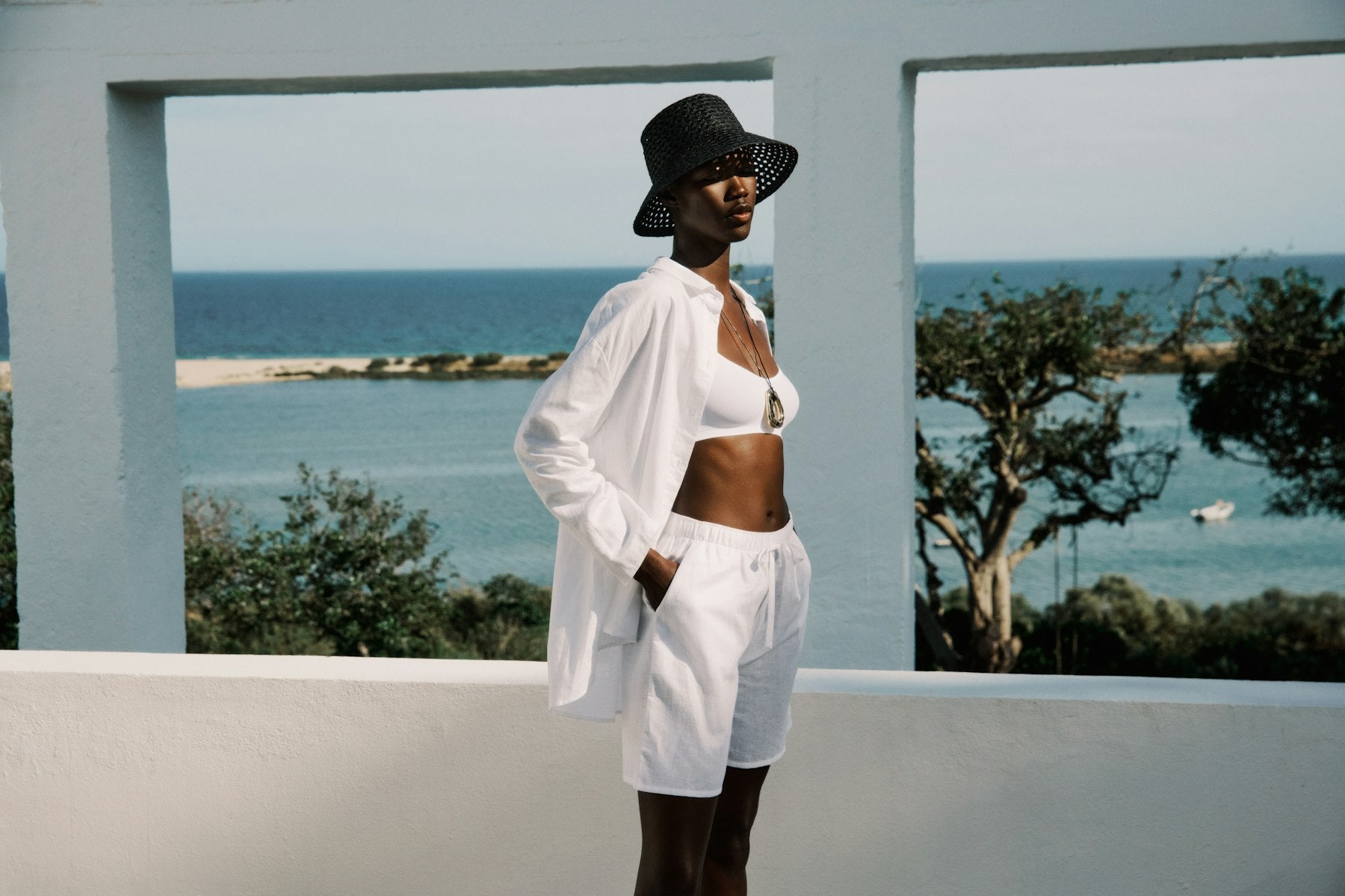 Person wearing a white outfit with a black hat standing in front of a scenic ocean view.