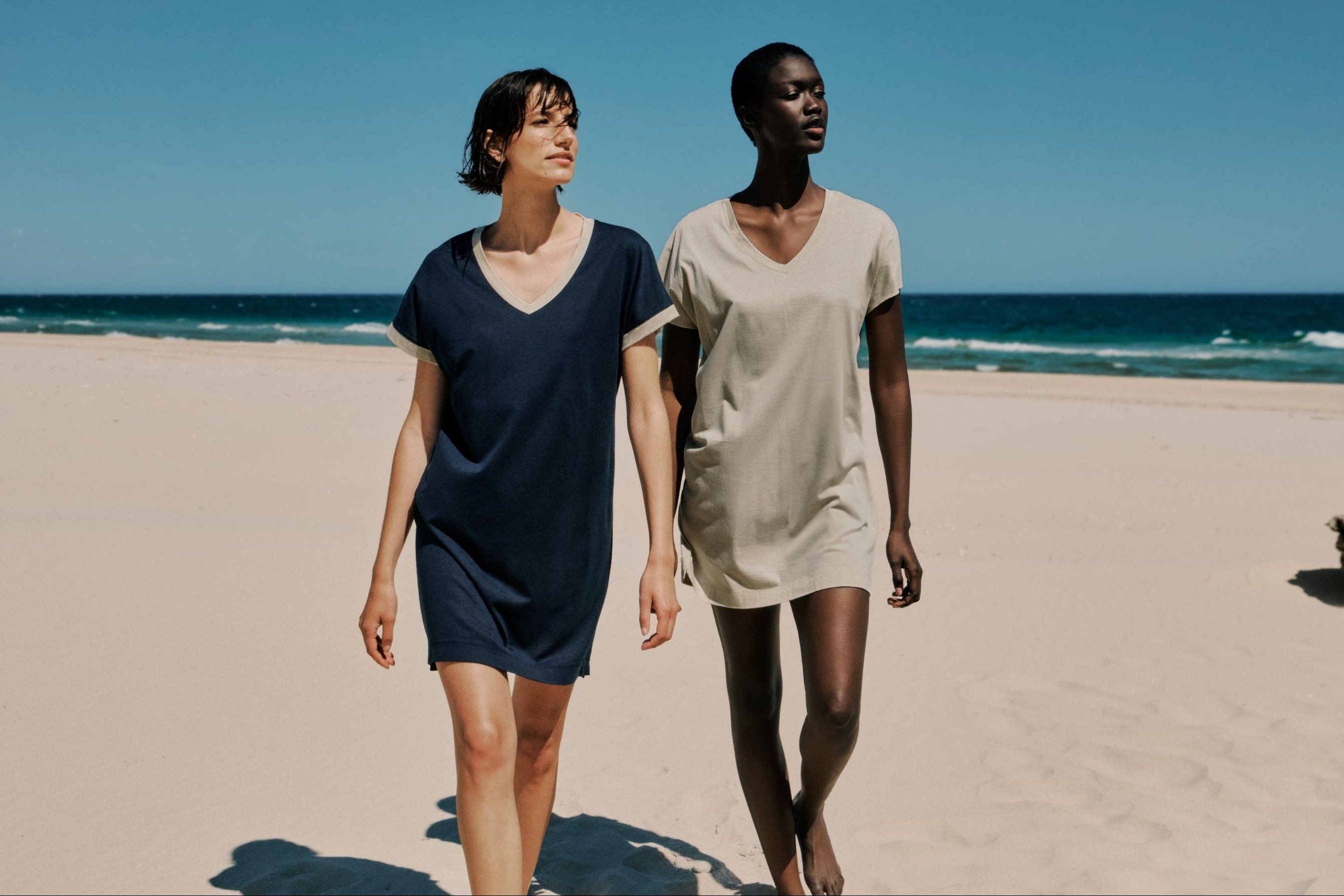 Two women walking on a sandy beach wearing simple V-neck bigshirts in navy blue and light beige, with the ocean and blue sky in the background.