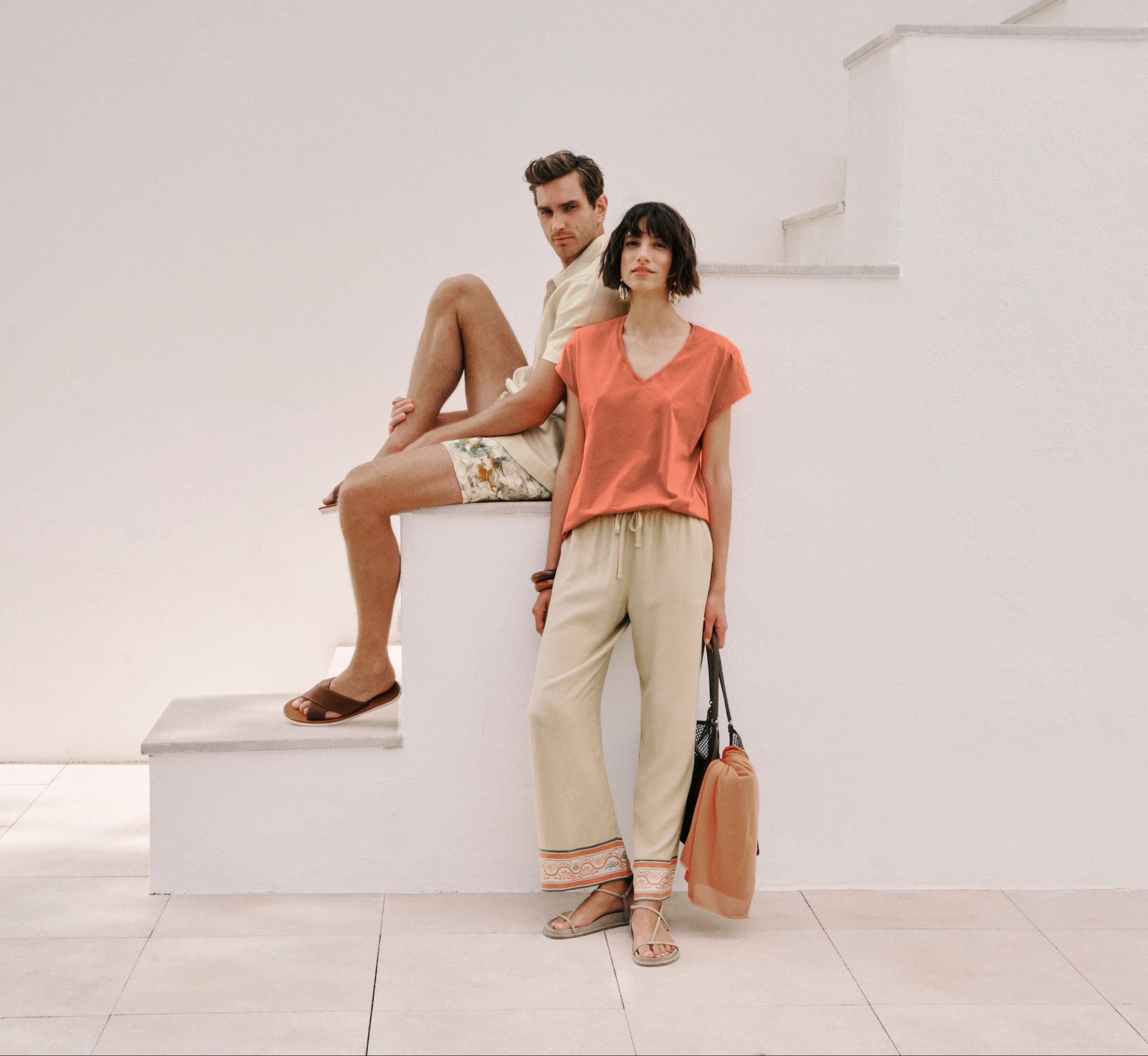 A man and a woman pose in a minimalist outdoor setting with steps. The woman wears a coral-orange short-sleeve top, beige wide-leg trousers, and holds a tan shoulder bag.