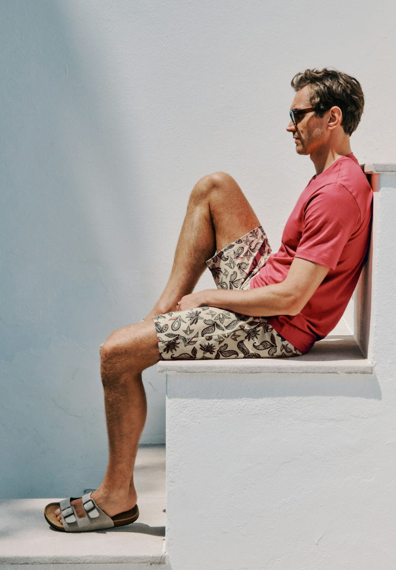 Man sitting on a white bench wearing a red shirt and patterned shorts.