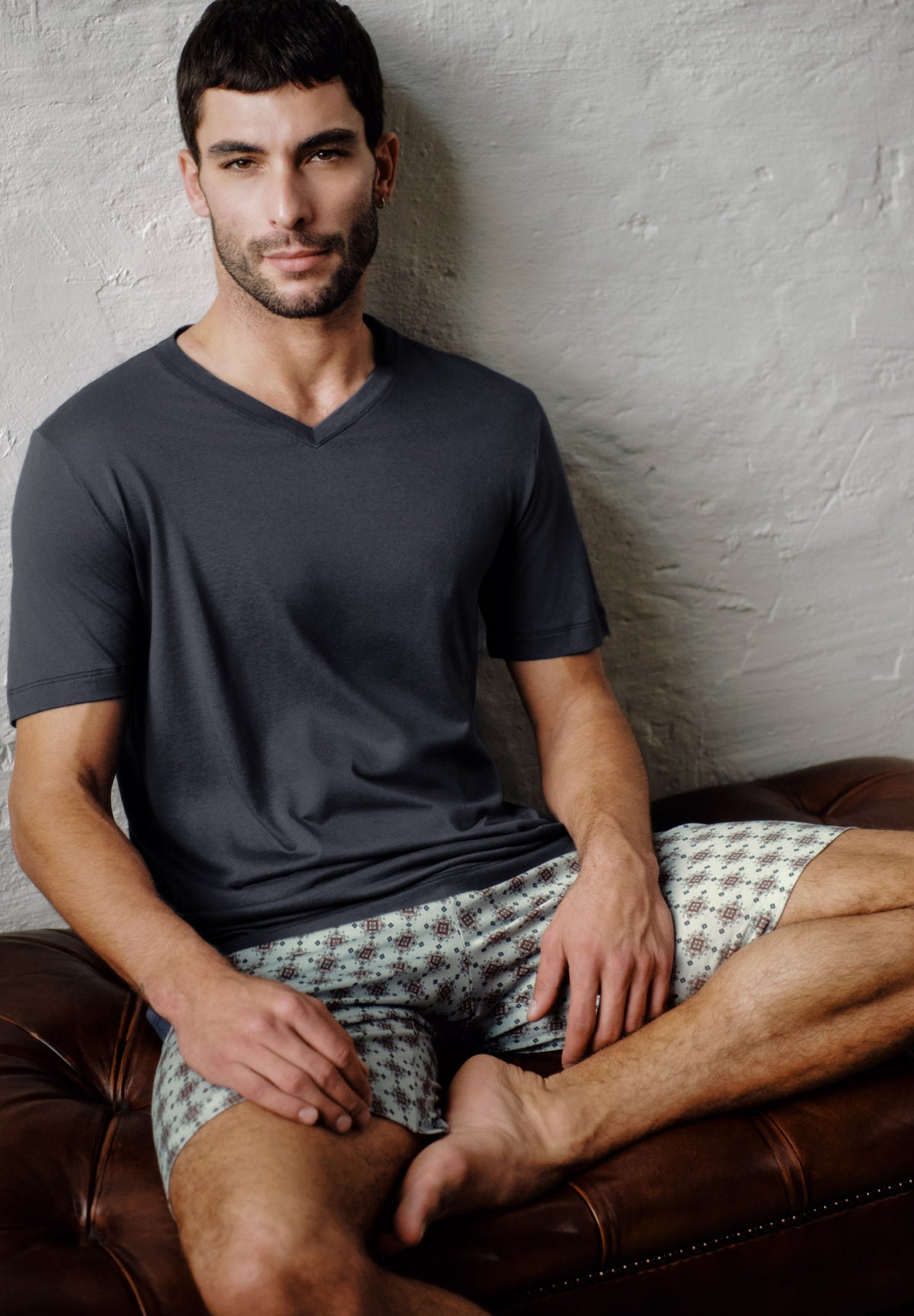 A man sitting on a brown leather couch against a textured light gray wall, wearing a dark charcoal gray v-neck t-shirt and patterned shorts.