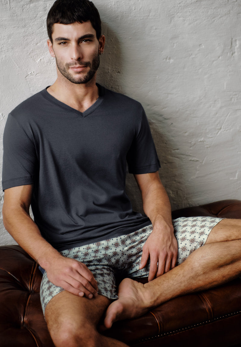 A man sitting on a brown leather couch against a textured light gray wall, wearing a dark charcoal gray v-neck t-shirt and patterned shorts.