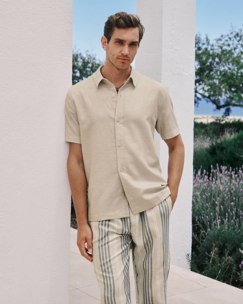 Man wearing a beige shirt and striped pants standing outdoors with greenery in the background