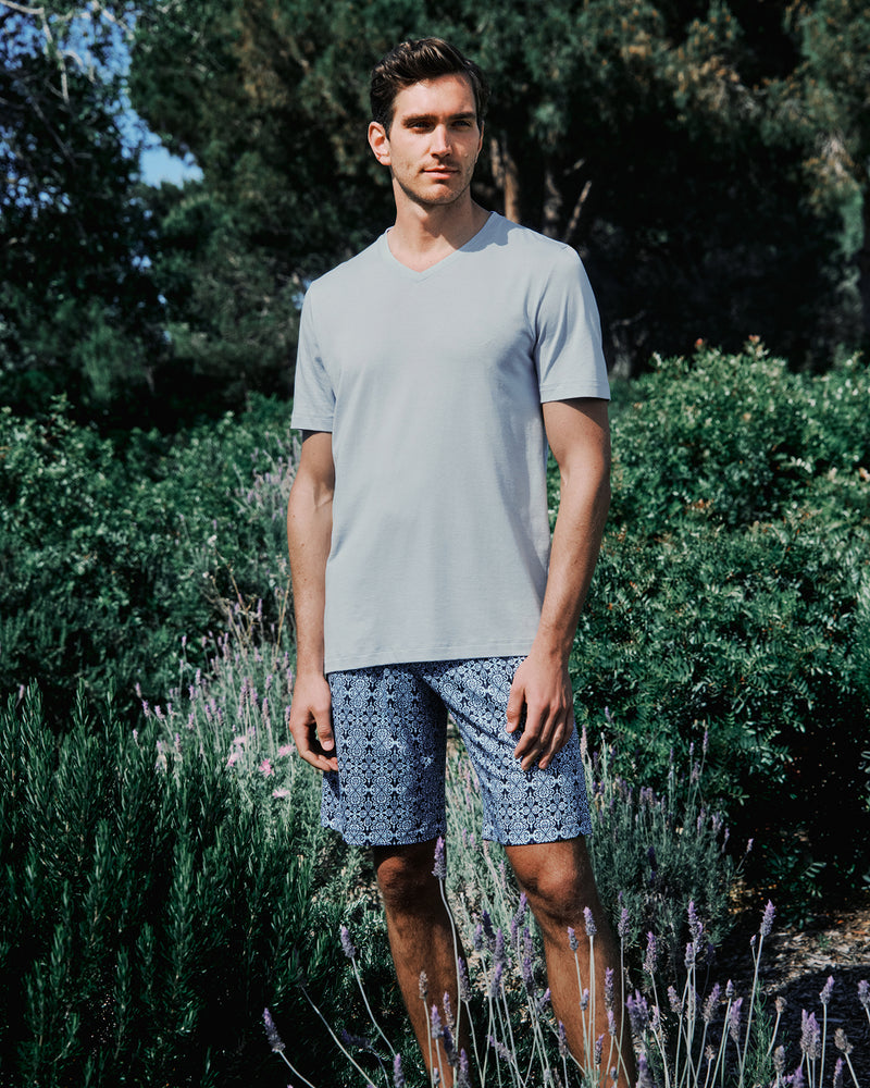 Man wearing a light blue t-shirt and patterned shorts standing in a garden with greenery and lavender.