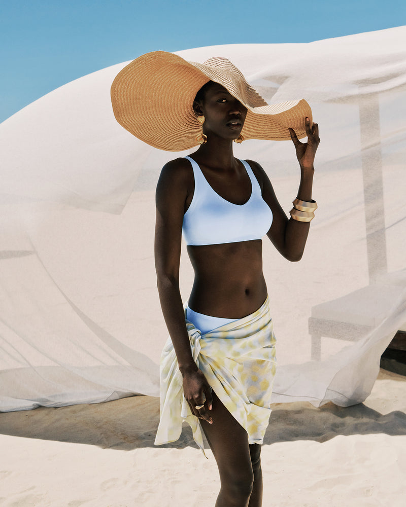 Woman in a blue bra and yellow skirt with a large straw hat on a beach.