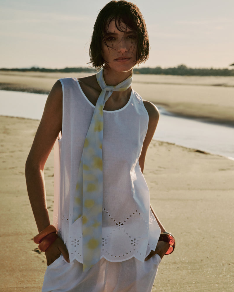 Woman standing on a beach wearing a white sleeveless top and a light-colored scarf.