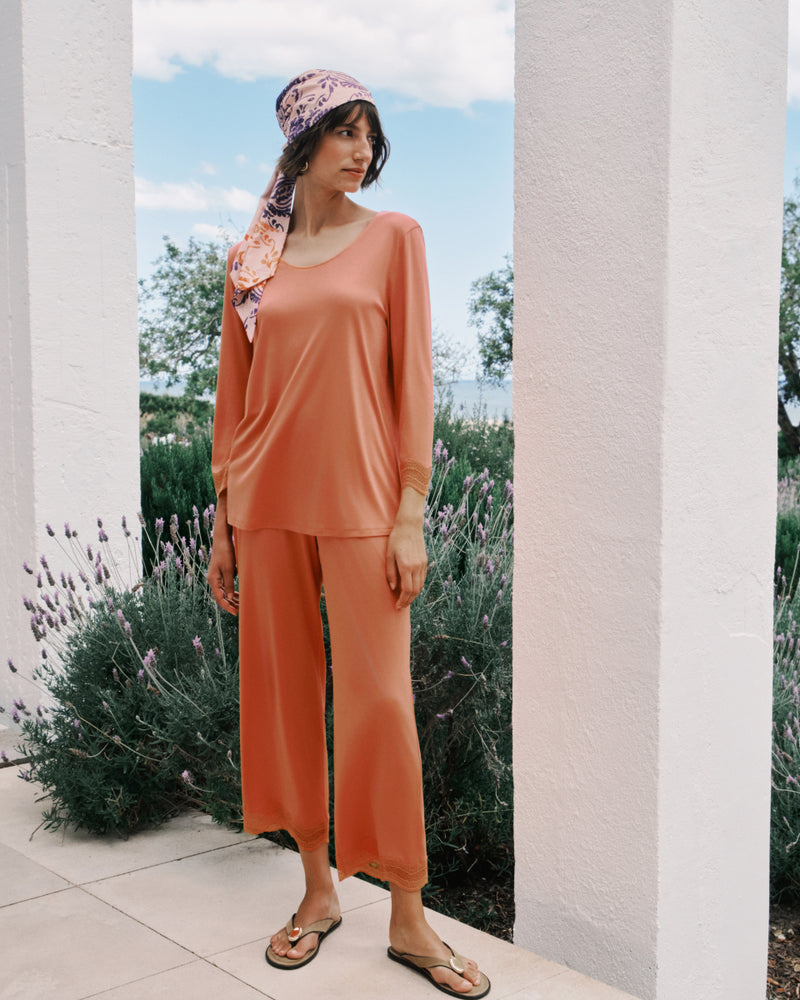 Woman in peach pajama set with floral headscarf standing outdoors near a white wall and lavender plants.