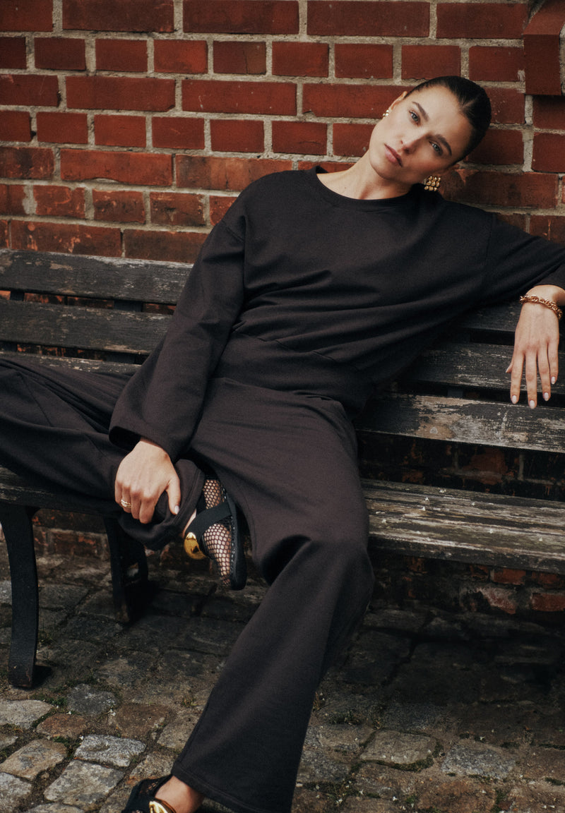 A woman wearing a black long-sleeve top and matching wide-leg pants sits on a weathered wooden bench against a red brick wall.