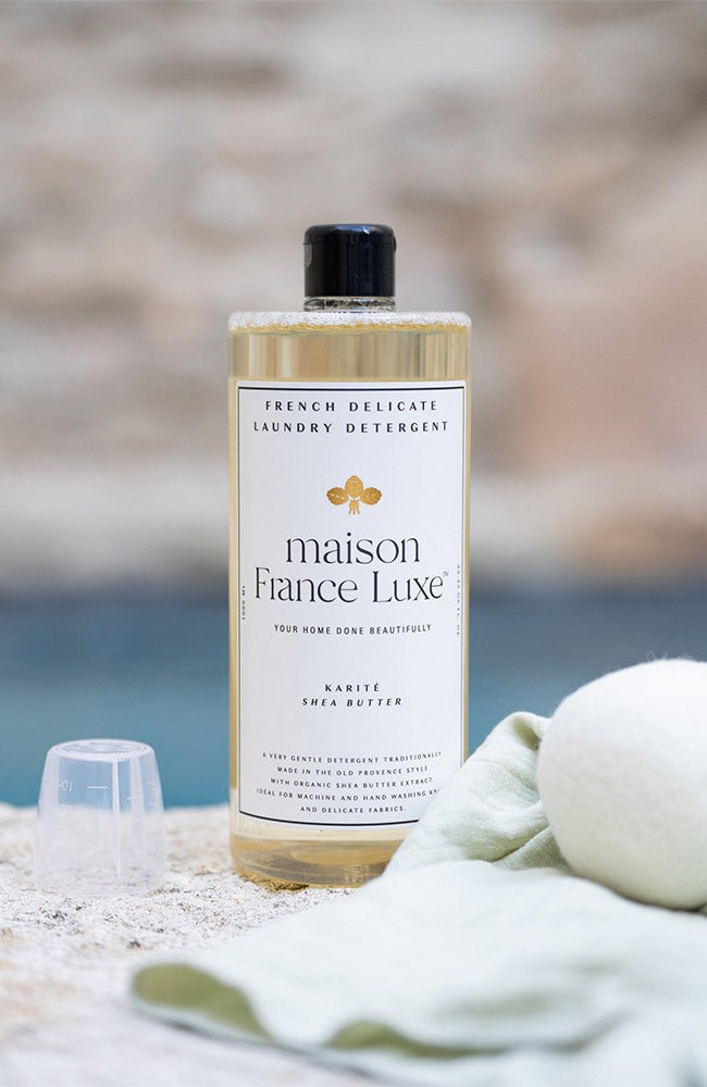 Bottle of Maison France Luxe laundry detergent on a beach with towels and sand.