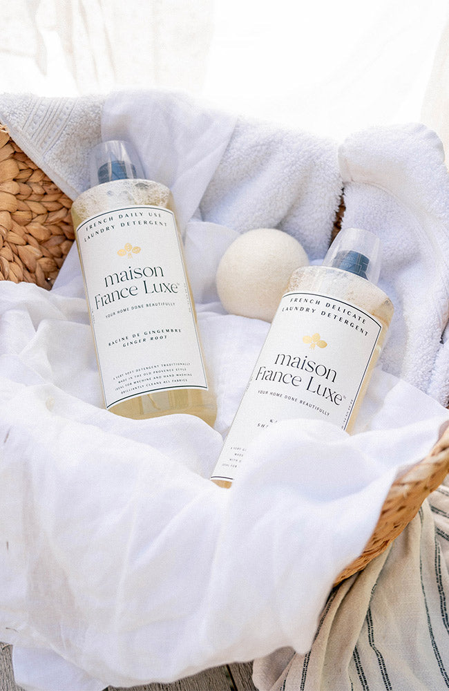 Two bottles of maison fance Luxe laundry detergent on a white blanket with a wooden basket.