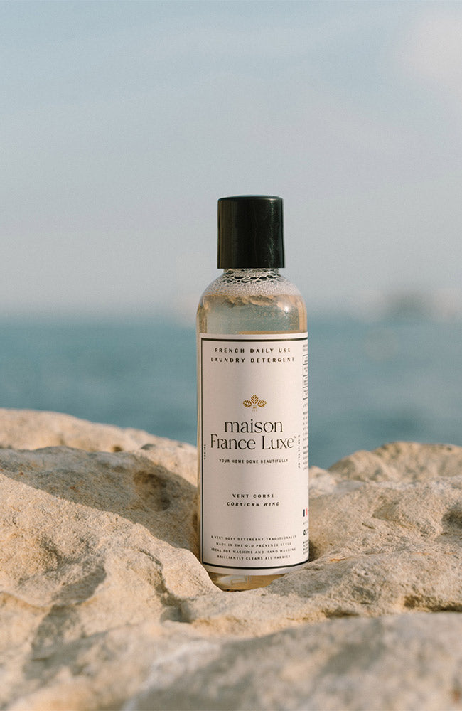 Bottle of Maison France Luxe laundry detergent on a rock with a blurred ocean background