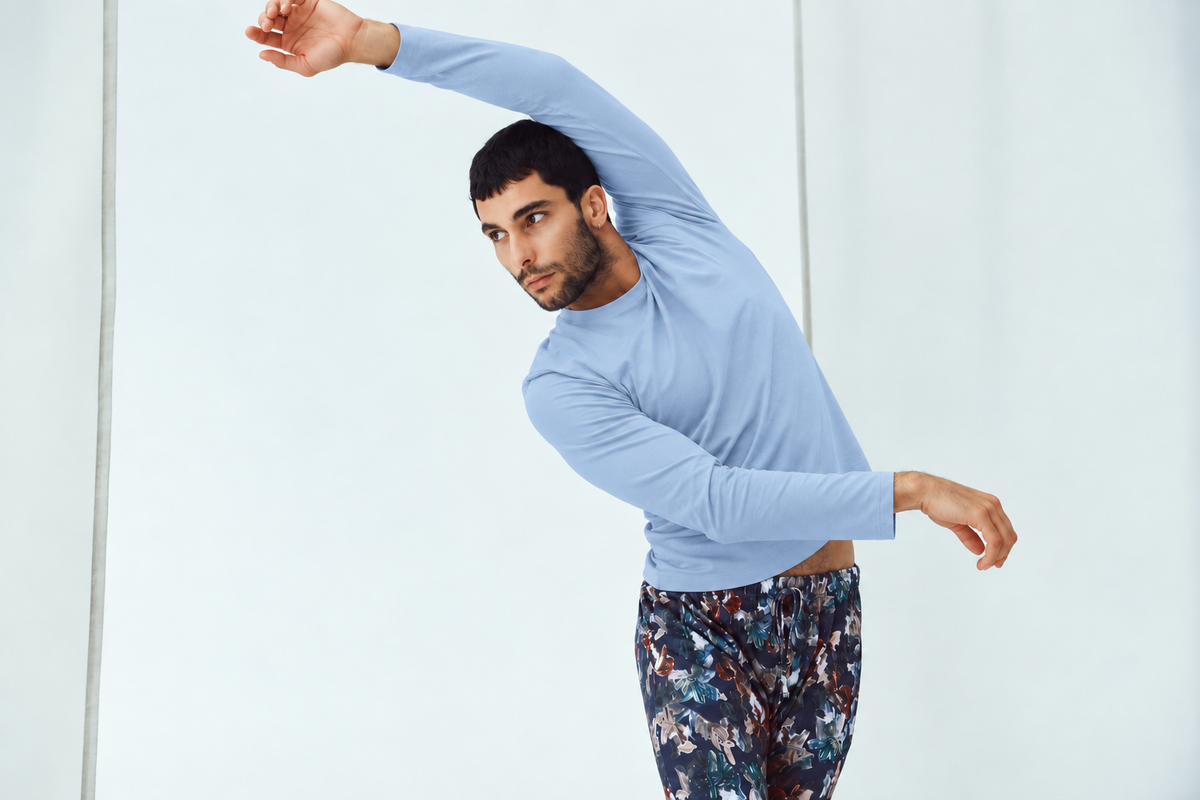 A man in a light blue long-sleeve top and dark floral-patterned pants poses in a side stretch against a clean white studio background.